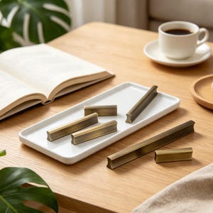 Puede incluir: Una colección de seis tiradores de armario de metal en tono latón con un diseño liso y alargado. Varios tiradores están dispuestos sobre una bandeja rectangular de cerámica blanca, mientras que otros dos descansan sobre la superficie de madera.