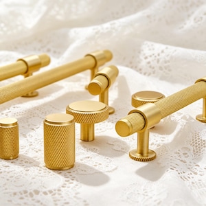 Op de afbeelding: Een collectie goudkleurige metalen kastbeslag, bestaande uit diverse cilindrische knoppen en lange staafgrepen. Elk onderdeel is voorzien van een opvallende, gekartelde kruisarcering op het oppervlak, wat zorgt voor een tactiele grip.