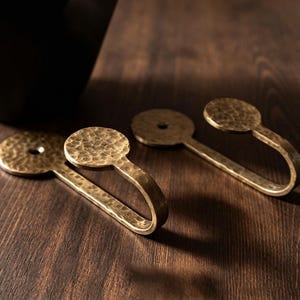 Può includere: Due ganci da parete in metallo color ottone con una finitura martellata e testurizzata. Ogni gancio presenta una piastra di montaggio circolare con un foro centrale per la vite e un braccio curvo a forma di J per appendere oggetti.