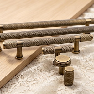 May include: A collection of antique brass cabinet hardware featuring a knurled texture. The set includes three long bar pulls of varying lengths, one T-bar handle, one round knob, and one cylindrical knob, all displaying a consistent metallic finish.