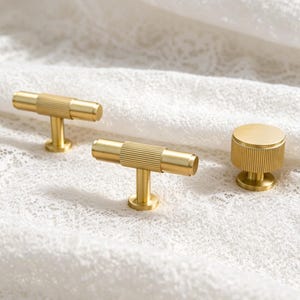 May include: Two gold-toned T-shaped cabinet pulls and one circular cabinet knob, all featuring a textured, ribbed surface design. These metal hardware pieces have a smooth, matte finish and a cylindrical central section with vertical grooves.