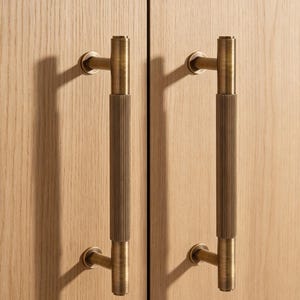 May include: Two vertical cabinet pulls made of antique brass with a cylindrical shape. Each handle features a central section with a fine, vertical ribbed texture, flanked by smooth, plain ends that connect to the cabinet door via two circular mounts.