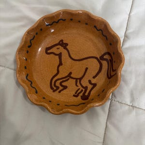 May include: A round, tan ceramic dish with a scalloped edge. The center features a dark brown line drawing of a horse in motion. The rim is decorated with a repeating pattern of small dark brown dots and wavy lines along the interior edge.
