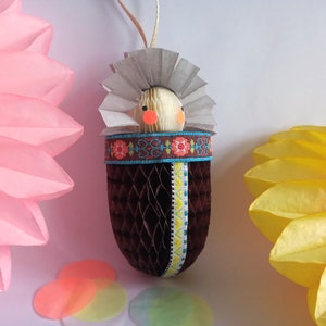 Op de afbeelding: Een bruin en wit papier-maché ornament in de vorm van een baby in een wieg. Het ornament heeft een witte papieren kroon en een bruin papieren lichaam met een geel en blauw lint. Het ornament is versierd met rode en blauwe bloemen.