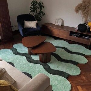 May include: Two nested wooden coffee tables with triangular tops and fluted cylindrical bases, positioned on a large, irregularly shaped area rug featuring a wavy pattern of light mint green and dark forest green tufted sections.