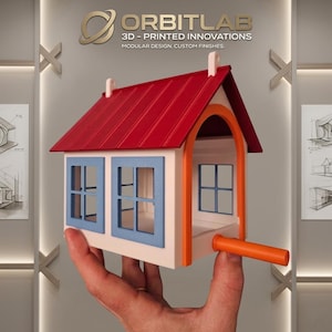 May include: A 3D-printed model house with a white base, a red roof, and blue window frames. The structure features an orange arched doorway and an orange cylindrical extension protruding from the side. The text ORBITLAB 3D-PRINTED INNOVATIONS MODULAR DESIGN, CUSTOM FINISHES is visible above the house.