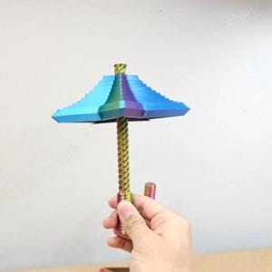 Könnte beinhalten: Ein metallisches, irisierendes Objekt in Regenschirmform mit einem gestuften, quadratischen Schirm, der von Blau zu Violett und Grün übergeht. Der zentrale Griff ist ein langer, spiralförmiger Stab mit regenbogenfarbenem Finish.