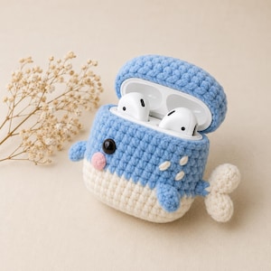 May include: A light blue and cream crochet case for wireless earbuds shaped like a whale. The case features a dark eye, a small pink cheek, white embroidered accents, and a tail. The lid is open, revealing white wireless earbuds inside.