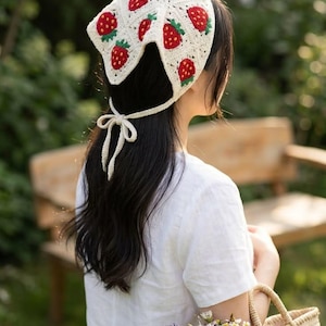 May include: A cream-colored crochet headscarf featuring a repeating pattern of red strawberries with green leaves. The headscarf is constructed from joined square motifs and includes thin crocheted ties that are knotted at the nape of the neck.