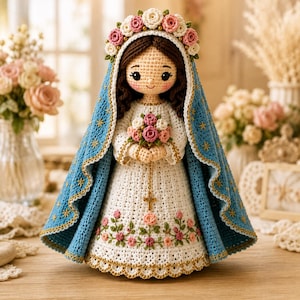 May include: A crocheted doll depicting a figure with dark brown hair, wearing a white dress with floral embroidery and a blue cloak adorned with gold star patterns. The figure wears a floral crown and holds a small bouquet with a gold rosary.