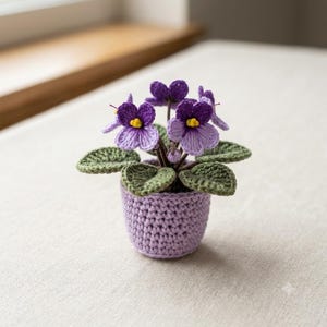 Könnte beinhalten: Eine gehäkelte Usambaraveilchen-Pflanze in einem kleinen, hellvioletten Garn-Topf. Die Pflanze hat dunkel- und hellviolette Blütenblätter mit leuchtend gelben Zentren, umgeben von strukturierten, dunkelgrünen gehäkelten Blättern aus Garn.