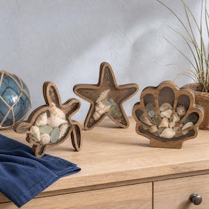 May include: Three wooden display frames shaped like a sea turtle, a starfish, and a seashell. Each frame features a clear front panel filled with various white seashells and small pieces of smooth sea glass, showcasing a natural coastal aesthetic.