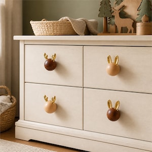 Peut inclure: Une commode en bois clair dotée de quatre tiroirs, chacun équipé d'un bouton sphérique. Deux boutons sont en bois brun foncé avec des bois de cerf dorés, et deux sont en bois clair avec des oreilles de lapin dorées fixées sur le dessus.
