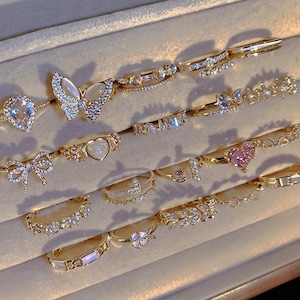 May include: A collection of gold-toned rings featuring various designs including a butterfly with iridescent wings, a pear-shaped stone, a heart-shaped stone, a bow, floral motifs, and bands accented with clear crystals and small white pearls.