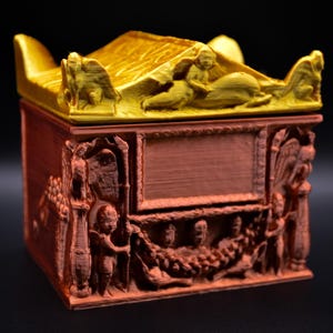 May include: A rectangular box with a terracotta-colored base and a bright gold lid. The lid features sculpted winged figures. The sides of the box display intricate relief carvings of cherubs, columns, and floral garlands surrounding a central panel.