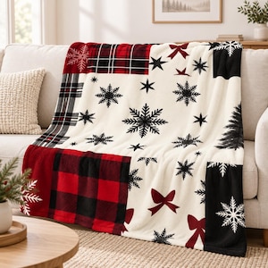 Puede incluir: Una manta de felpa con diseño de patchwork en secciones de color crema, rojo y negro. El patrón incluye copos de nieve negros, lazos rojos, cuadros rojos y negros, y la silueta negra de un pino sobre una tela suave y texturizada.