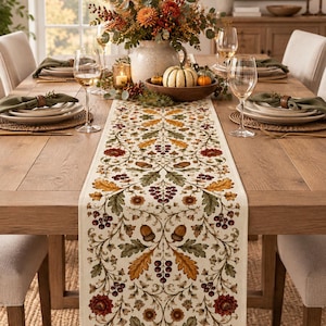 May include: A rectangular table runner featuring a cream-colored base with a repeating botanical pattern of dark red flowers, green and orange oak leaves, brown acorns, and clusters of small dark berries arranged in a symmetrical floral design.