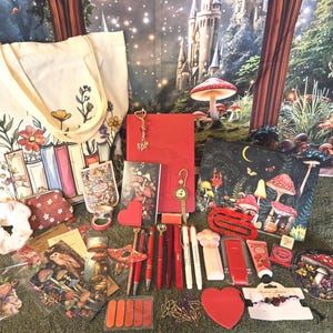Puede incluir: Una colección de papelería y accesorios que incluye una bolsa de tela crema con estampado de libros y flores, un cuaderno rojo, un estuche oscuro con diseño de bosque de setas, varios bolígrafos, artículos en forma de corazón y pegatinas.