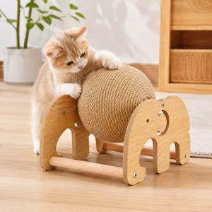 Puede incluir: Un gato de color marrón claro con patas blancas interactúa con una bola de cuerda de sisal beige montada sobre un marco de madera clara. El marco presenta dos paneles laterales con forma de siluetas de elefante y barras de soporte.