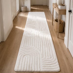 May include: A long, rectangular runner rug in a solid off-white color. The rug features a textured, raised pattern consisting of repeating arched and straight lines that create a geometric design across the entire surface of the floor covering.