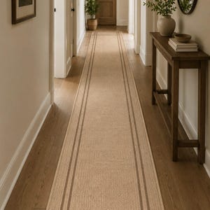 May include: A long, rectangular runner rug featuring a textured beige surface with dark brown border stripes and a cross-patterned design at the ends. The rug is positioned centrally on a wooden floor, extending down the length of a hallway.