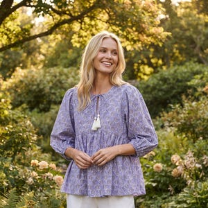 Puede incluir: Una blusa de color violeta claro con un estampado floral repetitivo en marrón y blanco, mangas largas con puños fruncidos y un cuello redondo con cierre de lazo terminado en dos borlas blancas, combinado con pantalones blancos holgados.