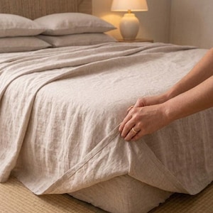 May include: A light beige linen bed sheet set featuring a flat sheet and matching pillowcases on a bed with a textured headboard. The fabric has a visible woven texture in a neutral oatmeal colour, draped over a mattress in a bedroom setting.