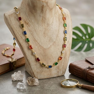 May include: A gold-tone chain necklace featuring a series of rectangular, multi-coloured gemstones in red, blue, green, and orange, interspersed with clear round stones. The necklace is displayed on a wooden bust, showcasing a vibrant, repeating pattern of colourful stones set in a metallic link chain design.