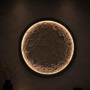 Può includere: Una lampada da parete circolare con una superficie testurizzata e craterizzata che imita la luna. Il pezzo è incorniciato da un bordo circolare scuro con retroilluminazione calda integrata che illumina il rilievo lunare. Il testo SELENE ART è visibile.