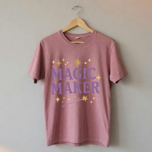 May include: A dusty rose short-sleeve t-shirt featuring the purple text MAGIC MAKER centered on the chest. The design is accented with yellow star icons and a small wand graphic. The shirt is displayed on a wooden hanger against a plain wall.