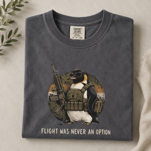 Peut inclure: Un t-shirt gris anthracite avec une illustration de manchot portant des lunettes de soleil et un équipement tactique, tenant un fusil. Le motif est sur fond circulaire avec le texte "FLIGHT WAS NEVER AN OPTION" imprimé sous l'image.