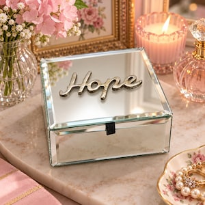 May include: A square mirrored jewelry box featuring a reflective glass surface and a silver-toned metal script word Hope attached to the lid. The box includes a small black fabric pull tab on the front edge for opening the hinged lid, showcasing a clean, polished design.
