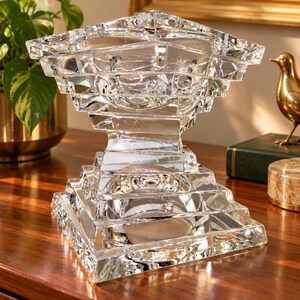 May include: A clear, faceted glass bowl with a square base and a wide, tiered rim. The object features geometric, stepped edges that create a multi-layered, prismatic effect, reflecting light through its transparent, heavy-duty glass structure.