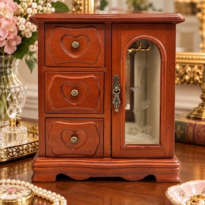 May include: A wooden jewelry armoire with a rich brown finish, featuring three stacked drawers with heart-shaped carvings and small brass knobs, alongside a side cabinet with a glass door etched with a floral design and a decorative metal handle.