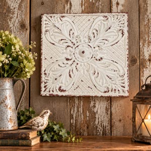 May include: A square metal wall tile featuring a white finish with distressed red edges. The surface displays an embossed floral pattern with a central circular medallion and a cross-hatched grid texture in the corners of the square panel.