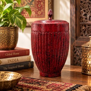 May include: A cylindrical red decorative vessel with a distressed finish and vertical fluted grooves. The container features a matching lid topped with a pointed finial. The surface displays a deep crimson hue with dark, weathered black accents.