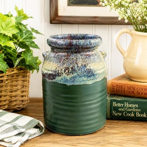 May include: A ceramic crock with a dark green ribbed base and a wide, flared rim featuring a mottled glaze of blue, brown, and cream. The vessel has two small side handles and a cylindrical shape, standing upright with a smooth, matte finish on the lower section.