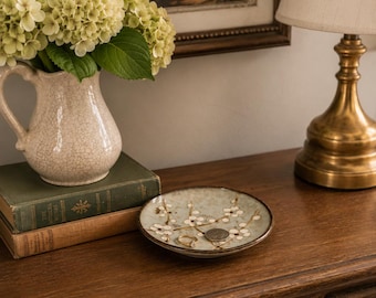 May include: A round ceramic trinket dish featuring a hand-painted floral pattern with white blossoms and brown branches on a light green background. The dish contains a gold-toned ring and a silver-toned coin resting on its surface.