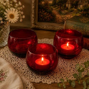 May include: Three round, deep red glass candle holders arranged on a circular lace doily. Two of the holders contain lit tea light candles with glowing orange flames, while the third holder is empty. The glass has a smooth, polished finish.