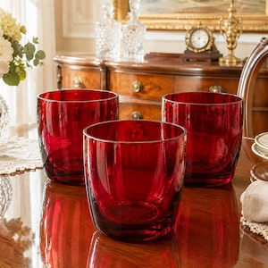 May include: Three deep red glass tumblers with a smooth, cylindrical shape and thick walls. The glasses are transparent, allowing light to pass through the vibrant crimson colour, and they feature a simple, unadorned design with a flat base.