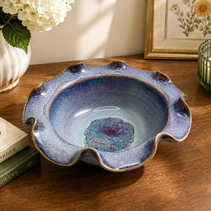 May include: A ceramic bowl with a wide, ruffled edge featuring a gradient glaze that transitions from deep indigo and violet at the rim to a lighter, mottled blue and teal in the center, showcasing a textured, crystalline finish throughout.