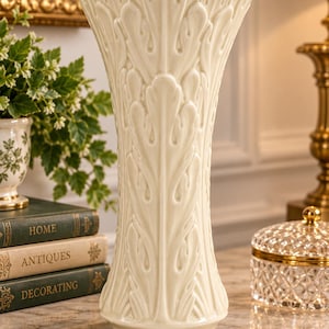 May include: A tall, cream-colored ceramic vase featuring a prominent, raised vertical leaf pattern that extends from the base to the scalloped, flared rim. The surface has a smooth, glossy finish that reflects light across its textured exterior.