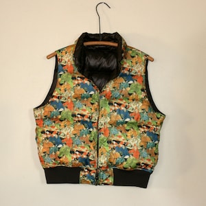 May include: A black puffer vest with a colorful print of people. The vest has a zipper closure and a black band at the bottom.