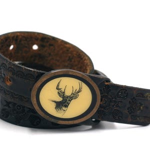 May include: A brown leather belt with an ornate floral design and a round buckle featuring a white deer silhouette.