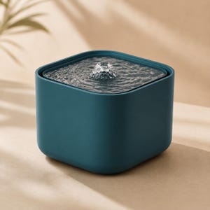 May include: A square, dark teal pet water fountain with rounded corners. The top surface features a central nozzle where water bubbles upward, creating concentric ripples across the surface of the clear water contained within the basin.