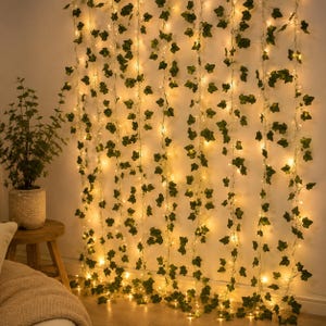 Peut inclure: Une décoration murale composée de multiples brins verticaux de feuilles de lierre artificielles vertes entrelacées avec des guirlandes lumineuses LED blanc chaud. Les brins pendent parallèlement, créant un effet de rideau végétal lumineux.