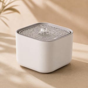 May include: A square-shaped pet water fountain with a matte white exterior and a grey top rim. The device features a central nozzle that creates a gentle, bubbling water flow on the surface, providing a continuous stream for hydration needs.