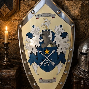 May include: A pointed heater shield with a cream-colored background and a metallic silver border featuring rivets. The center displays a blue crest with a yellow star, crossed arrows, a knight's helmet, a unicorn, and the text Trabue and France.