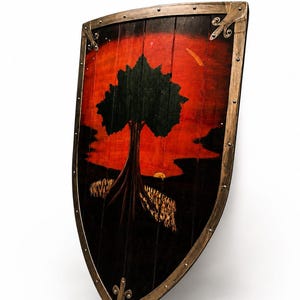 May include: A wooden heater shield featuring a painted scene of a black tree silhouette against a vibrant red and orange sunset background. The shield has a metallic, weathered-look border secured with visible rivets along the outer edges.
