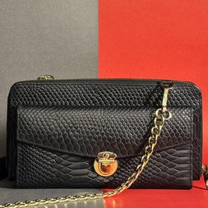 Bolsa transversal tipo clutch em couro bovino preto com estampa de cobra e carteira com corrente dourada.